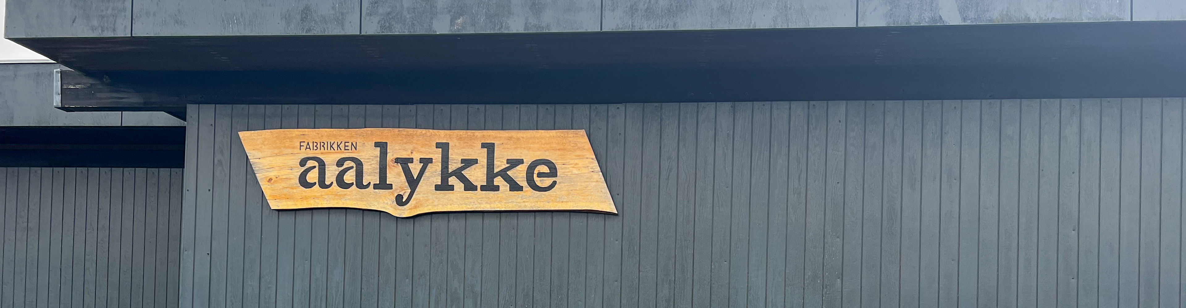 Sort træbygning med et træskilt på, hvor der står Fabrikken Aalykke på